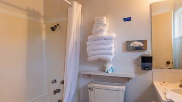 A bathroom with a shower, toilet, sink, and a stack of towels on a shelf.