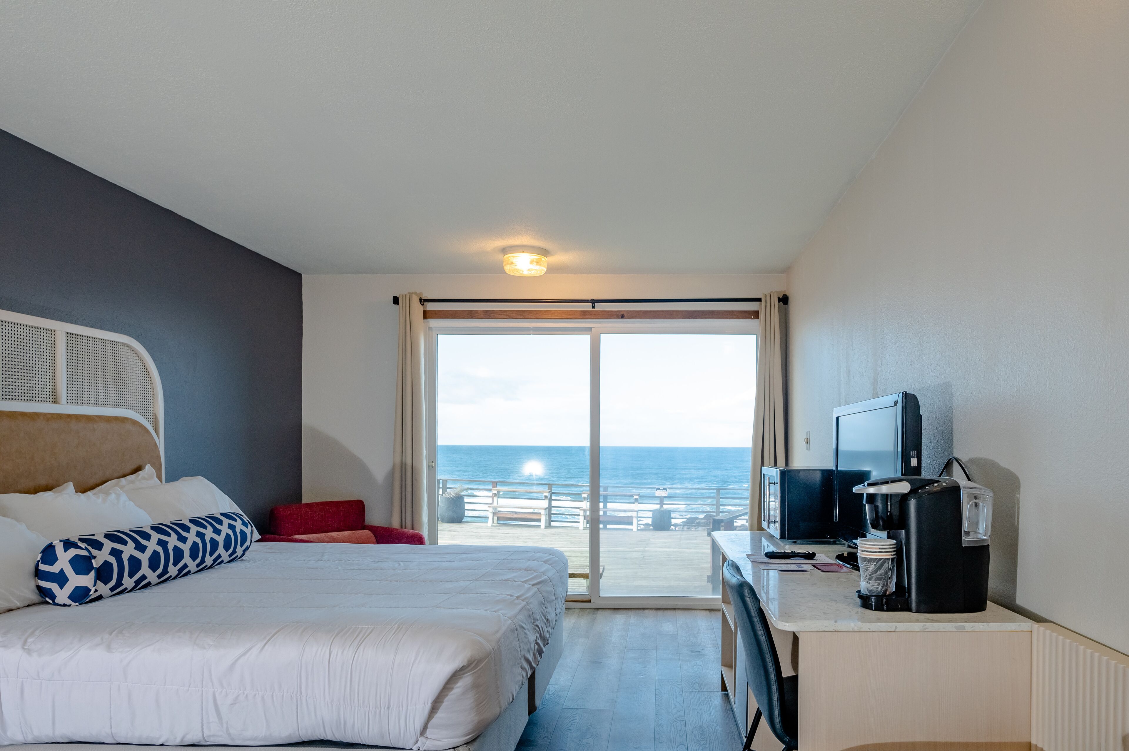 A hotel room with a bed, a desk with a coffee maker, a television, and a view of the ocean through a sliding door.