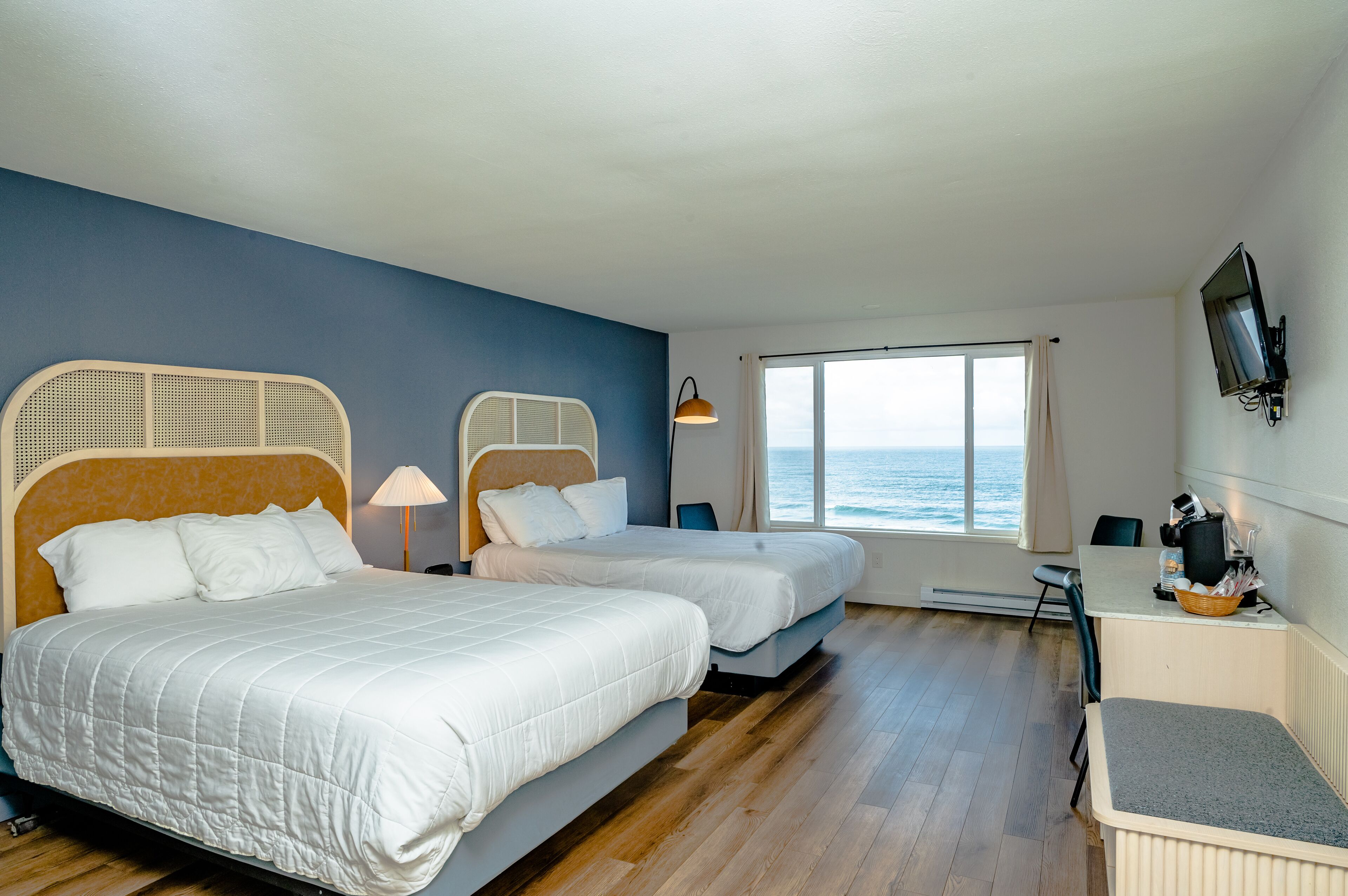 A hotel room with two beds, a desk, a chair, a TV, and a view of the ocean.