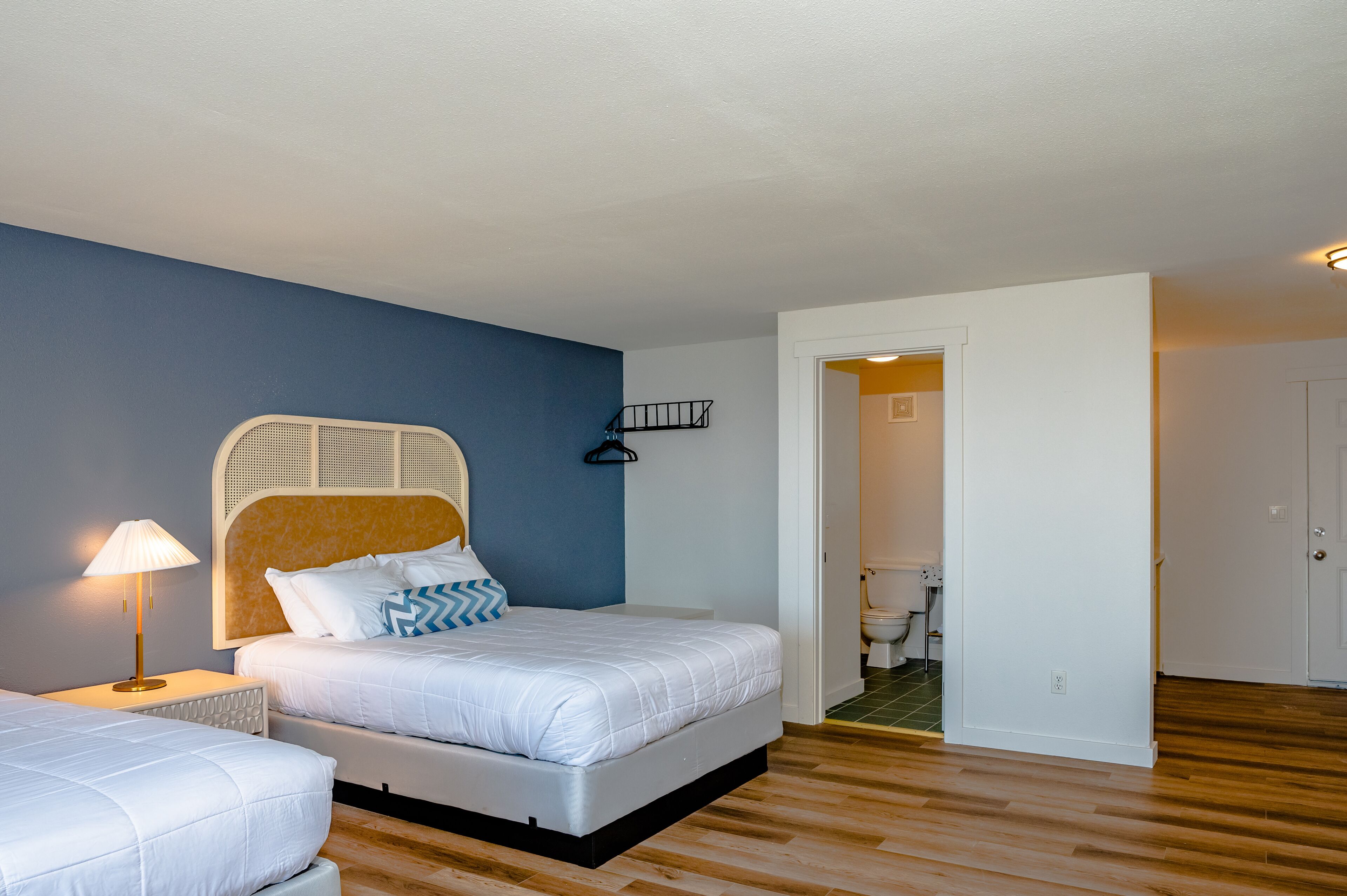 A bedroom with a bed, bedside table, and lamp. There is a door leading to another room.