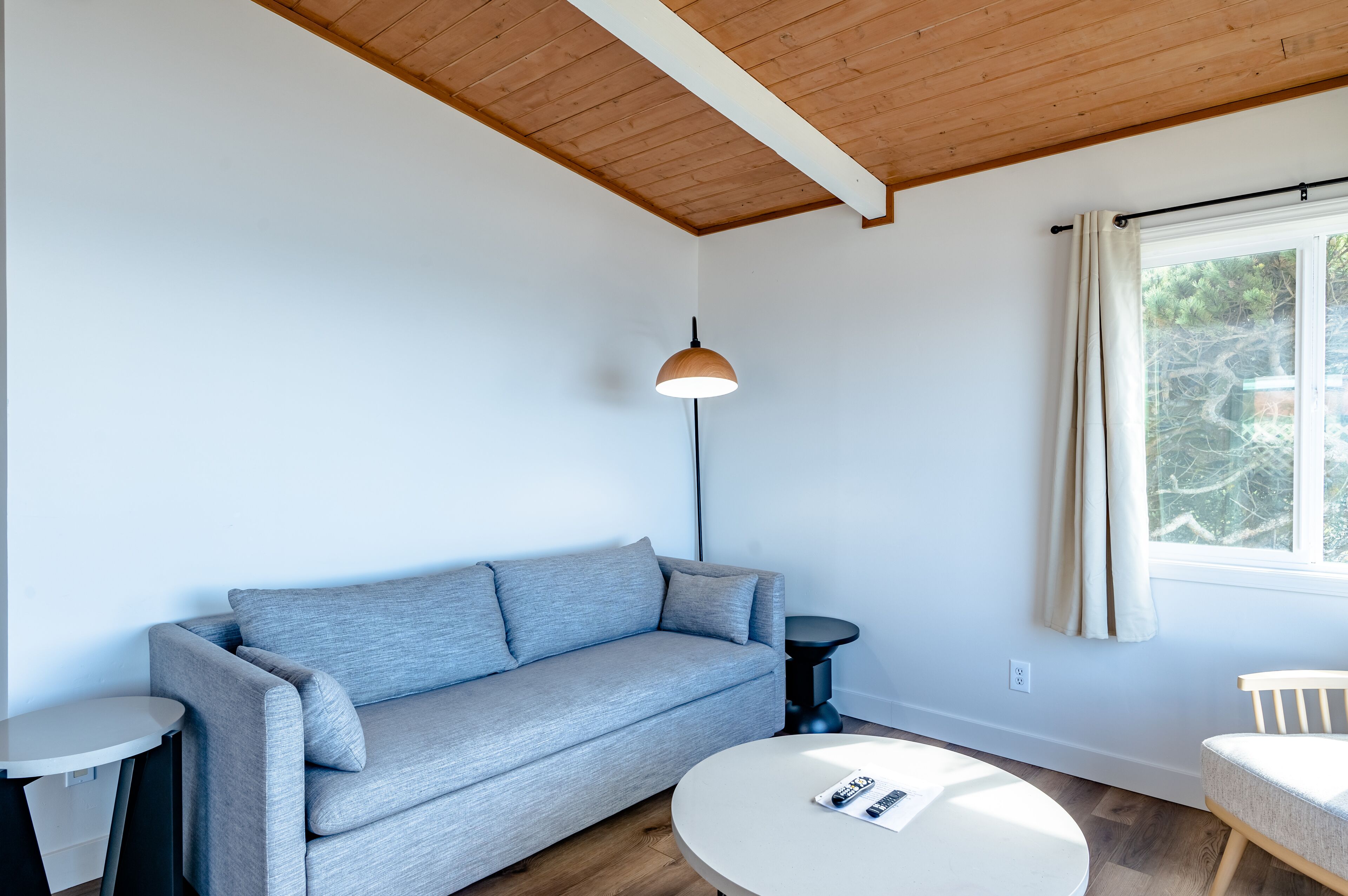 Una sala de estar moderna con un sofá gris, una mesa de centro redonda, una lámpara de pie y una ventana con cortinas.