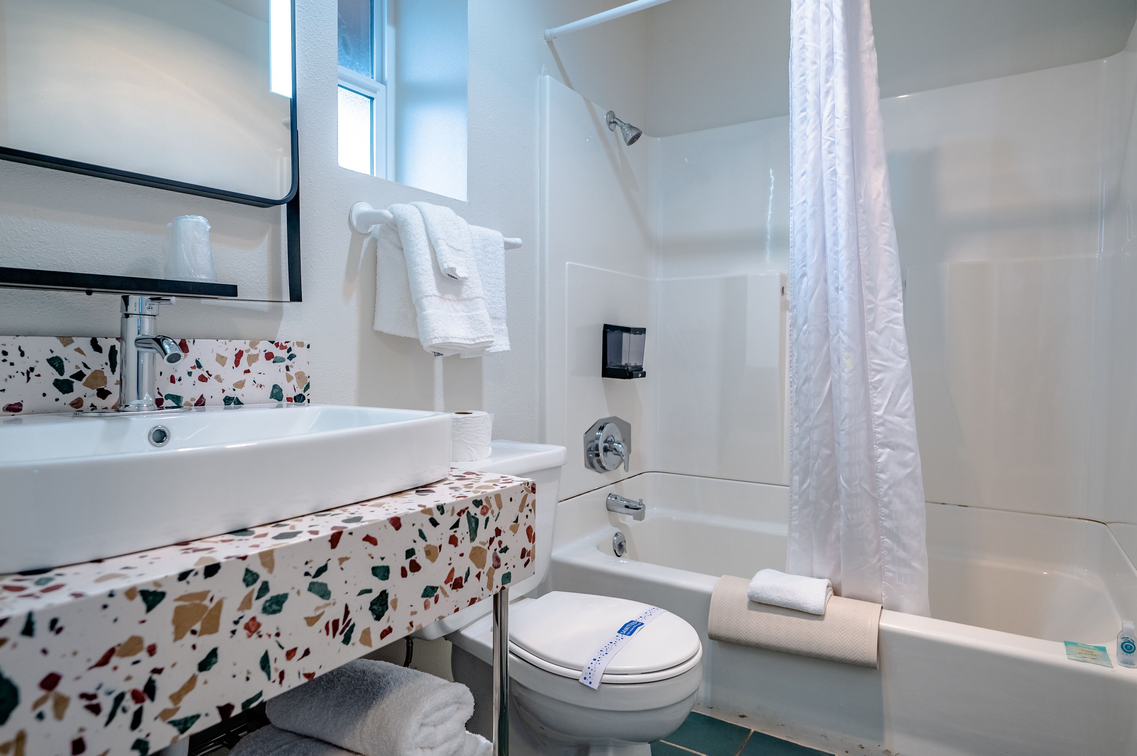 Une salle de bain avec un lavabo blanc, un miroir et des toilettes, un espace douche avec un rideau blanc, et un meuble-lavabo à motifs de terrazzo.