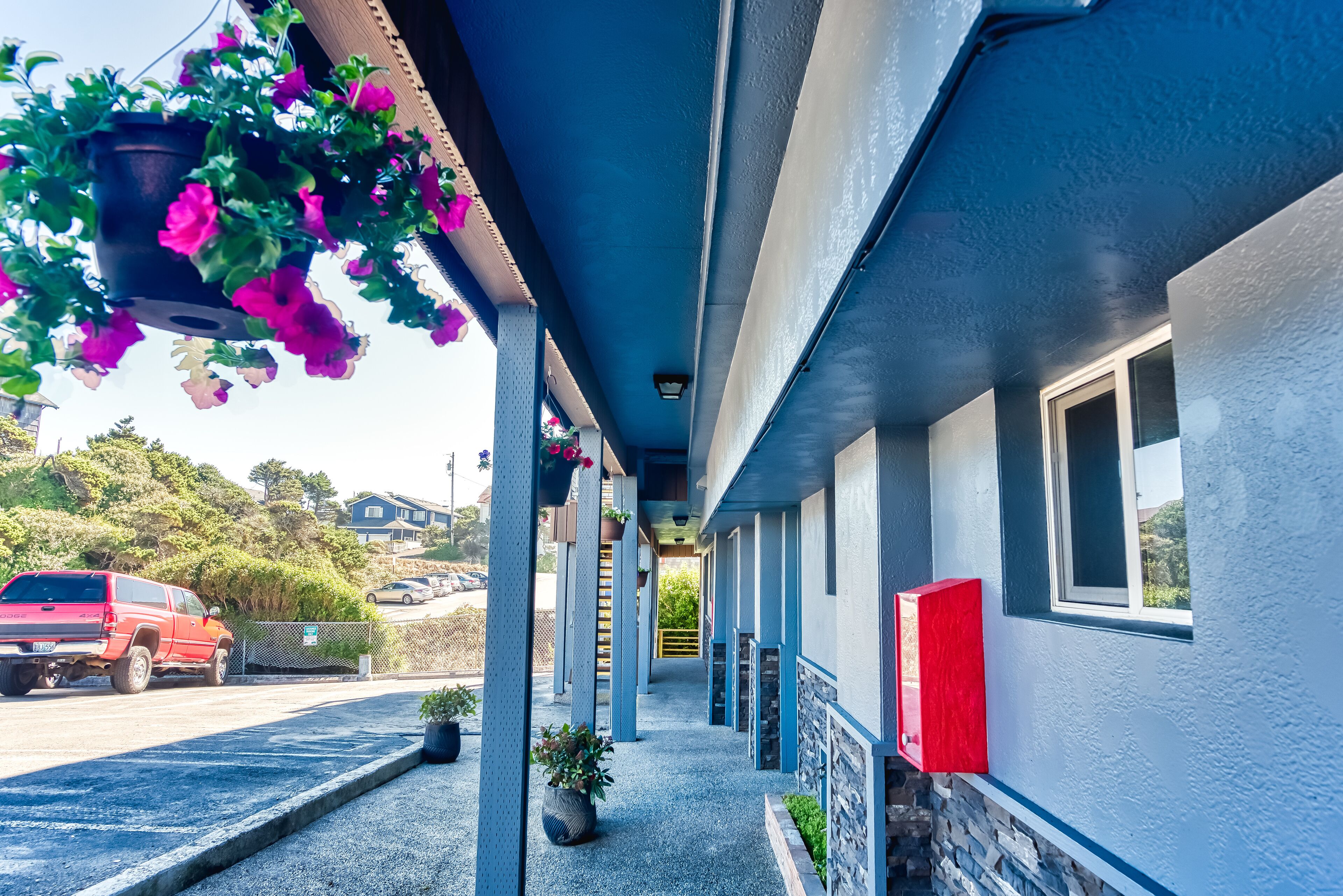 A covered walkway with hanging flower baskets, a red vehicle parked outside, and a building with a stone base and blue walls.
