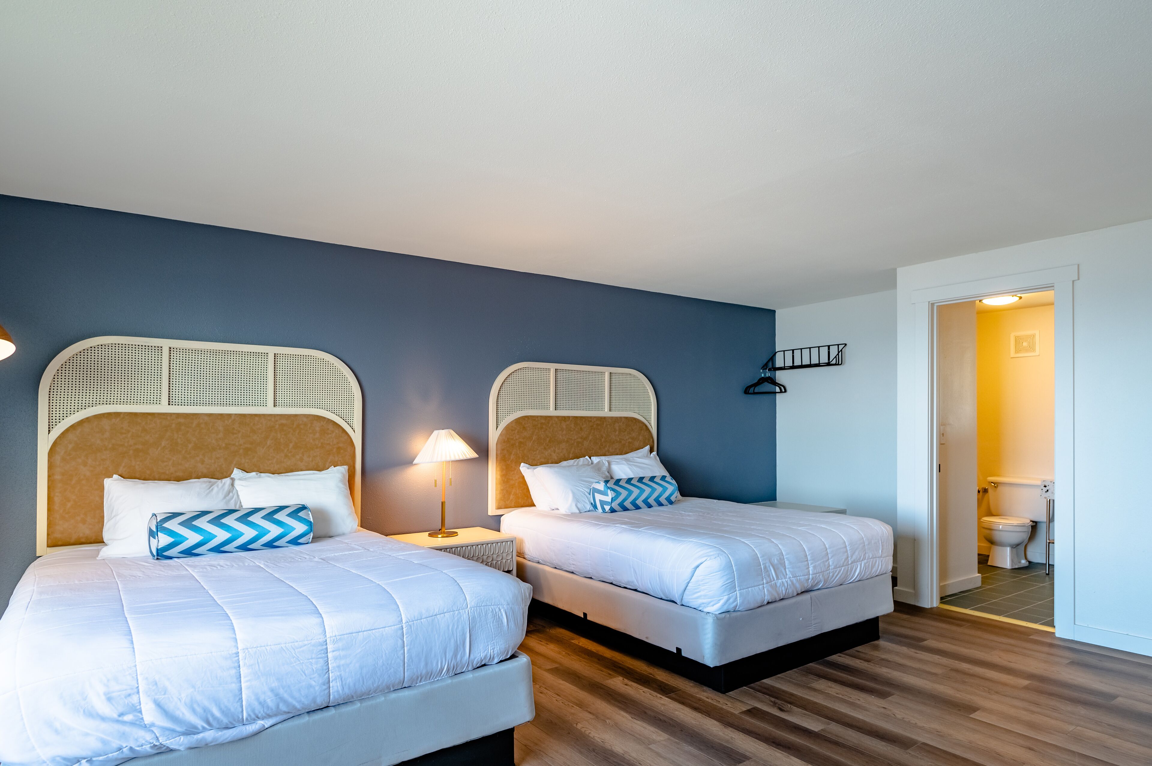 Quarto de hotel com duas camas, piso de madeira, parede azul e porta branca que dá para um banheiro.