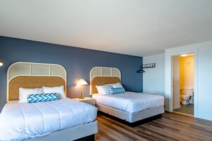 Quarto de hotel com duas camas, piso de madeira, parede azul e porta branca que dá para um banheiro.