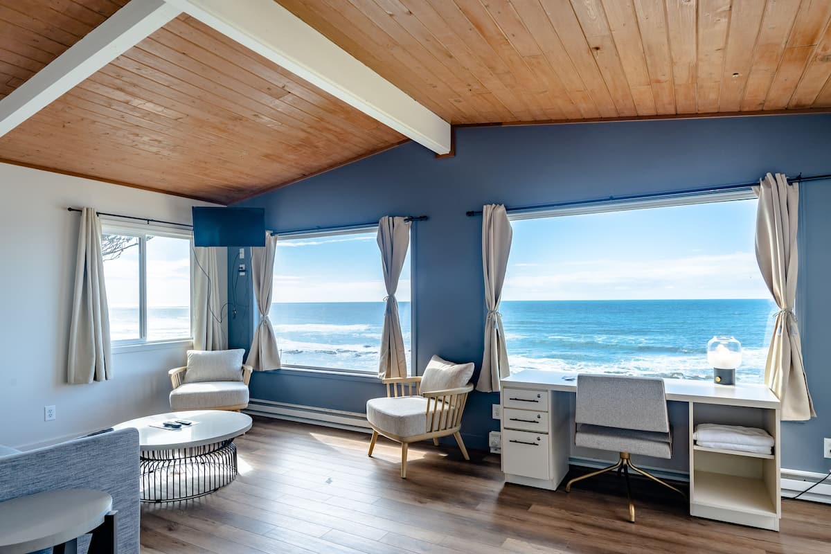 Een kamer met een houten plafond, grote ramen voorzien van gordijnen, een ronde tafel, een stoel, een bureau en uitzicht op de oceaan.
