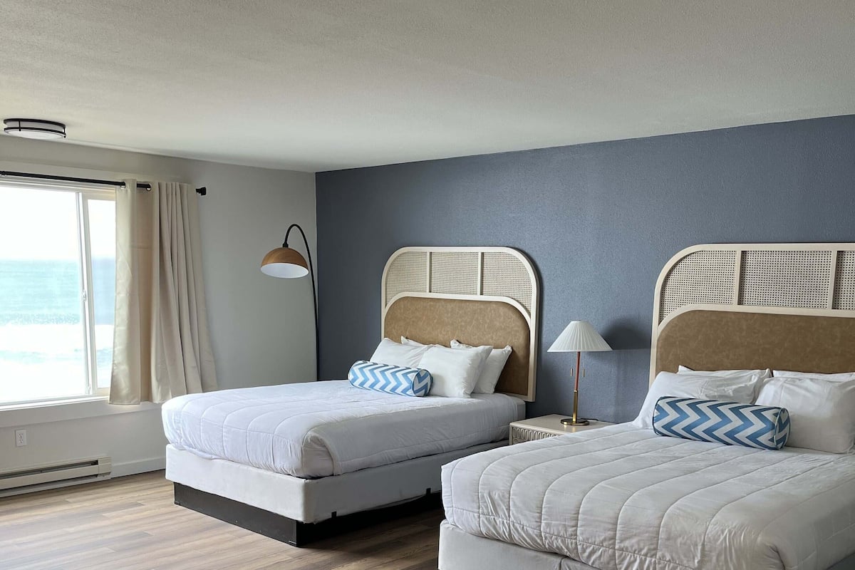 Een hotelkamer met twee bedden, een groot raam met gordijnen, een staande lamp en uitzicht op de oceaan.
