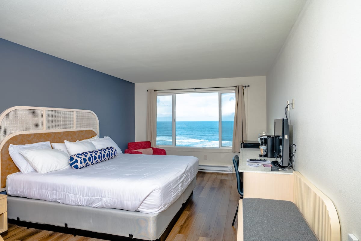 Een slaapkamer met een bed, een rode stoel, een bureau met een computer en een raam met uitzicht op de oceaan.