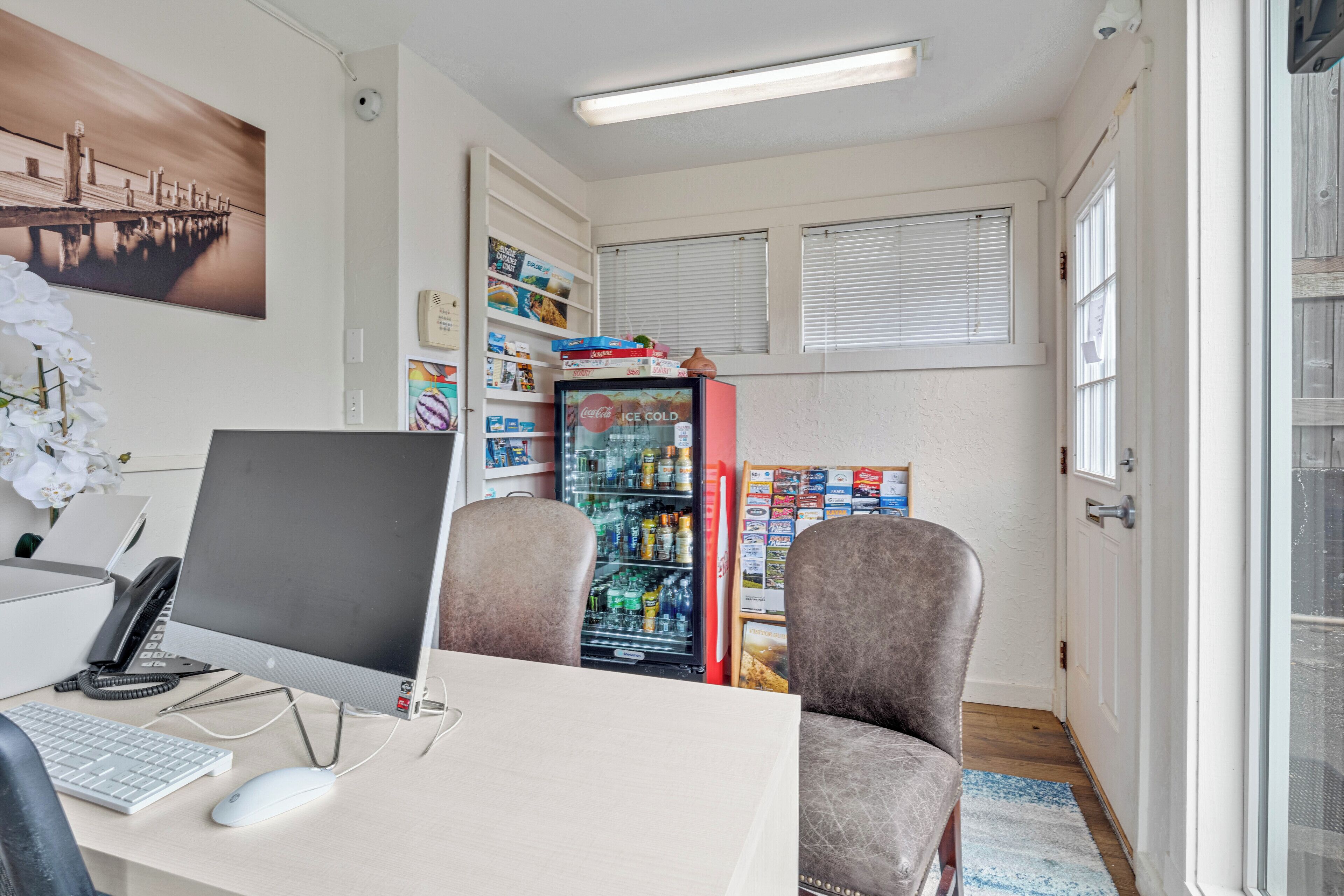 A room with a desk, computer, two chairs, a refrigerator, and a window with blinds.