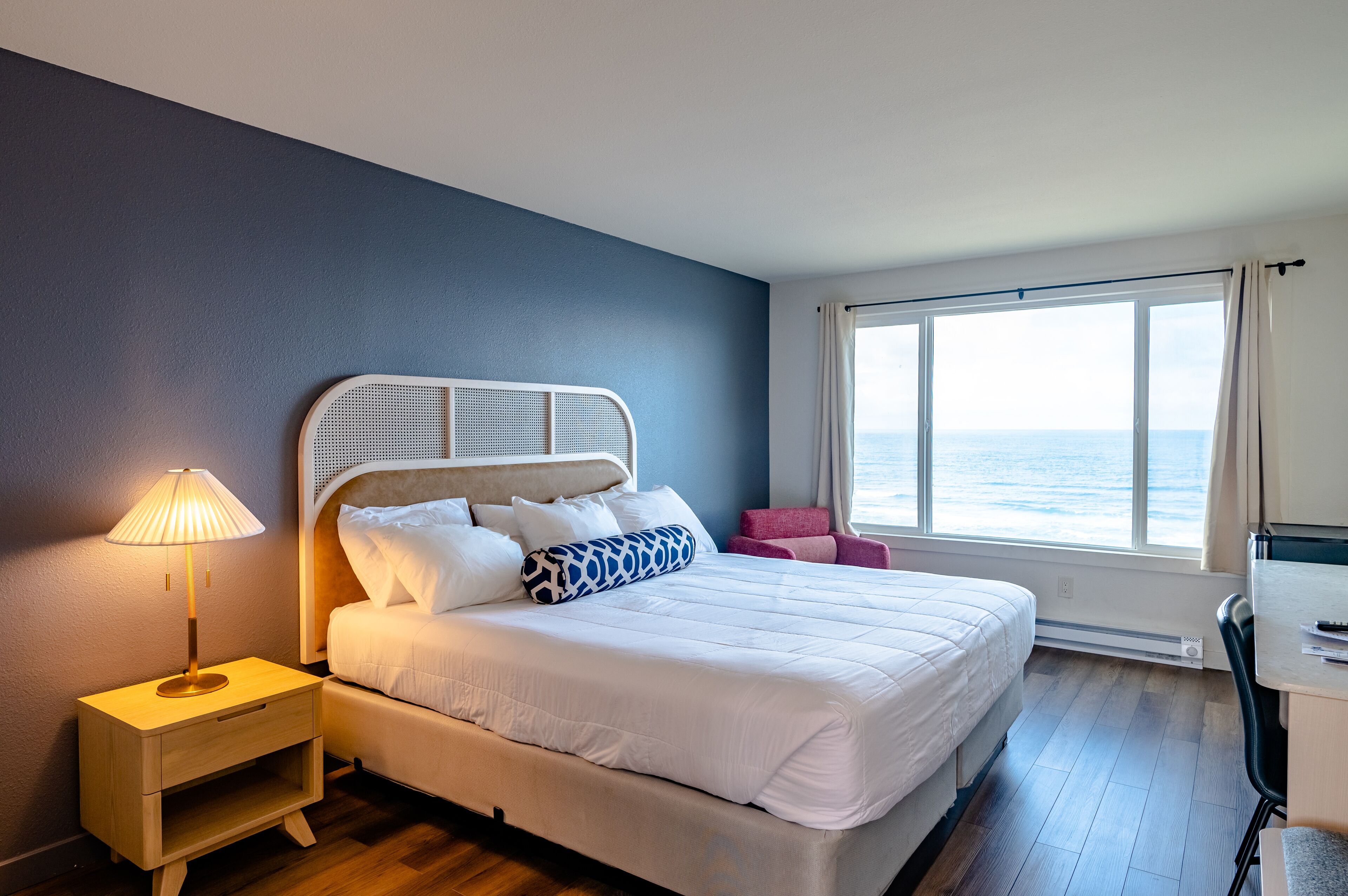 A neatly made bed with white bedding, a bedside lamp, and a view of the sea through a large window.