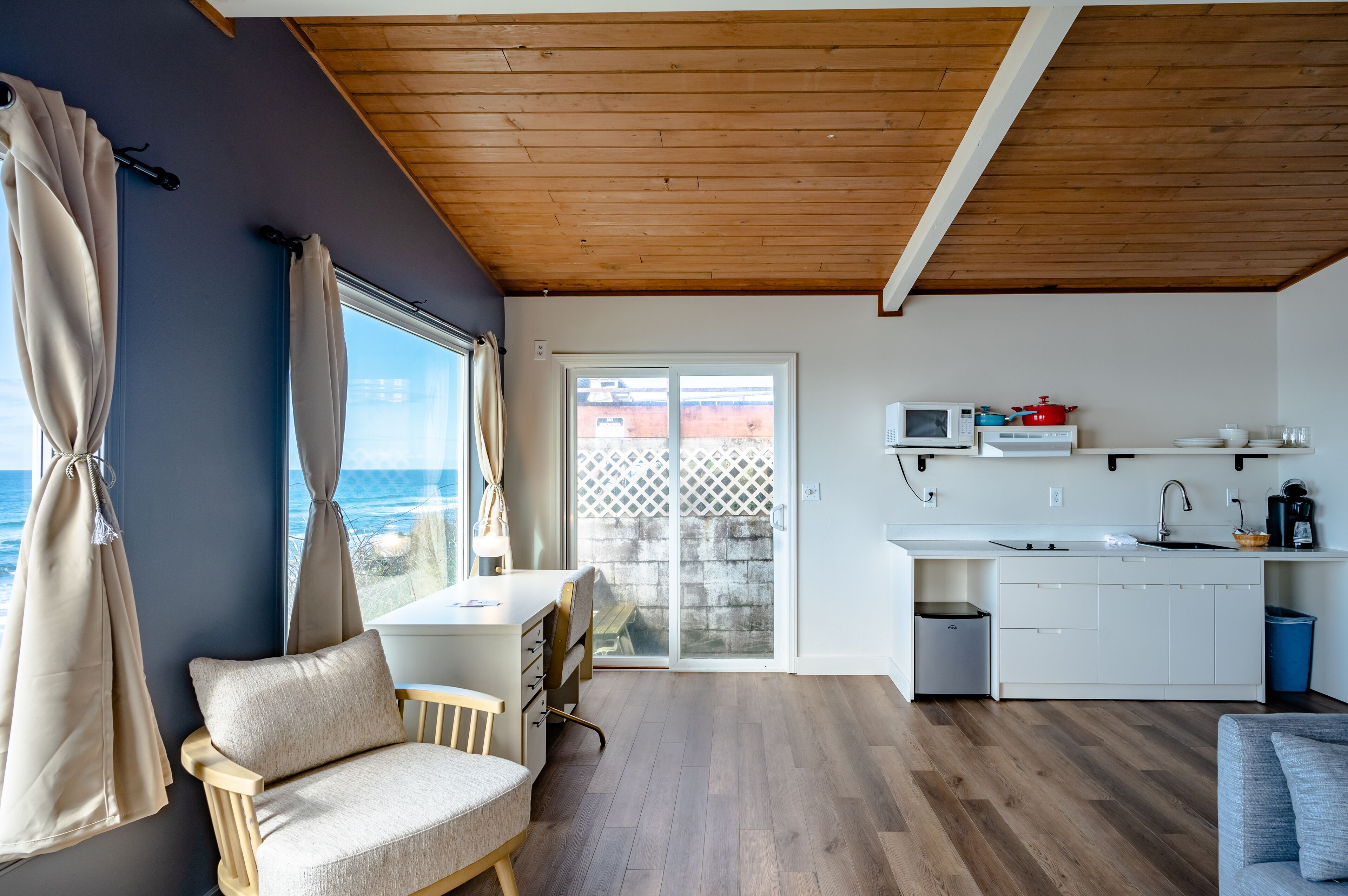 A modern kitchen with white cabinets, a microwave, and a coffee maker. There is a dining area with a table and chairs, and a view of the ocean through large windows.