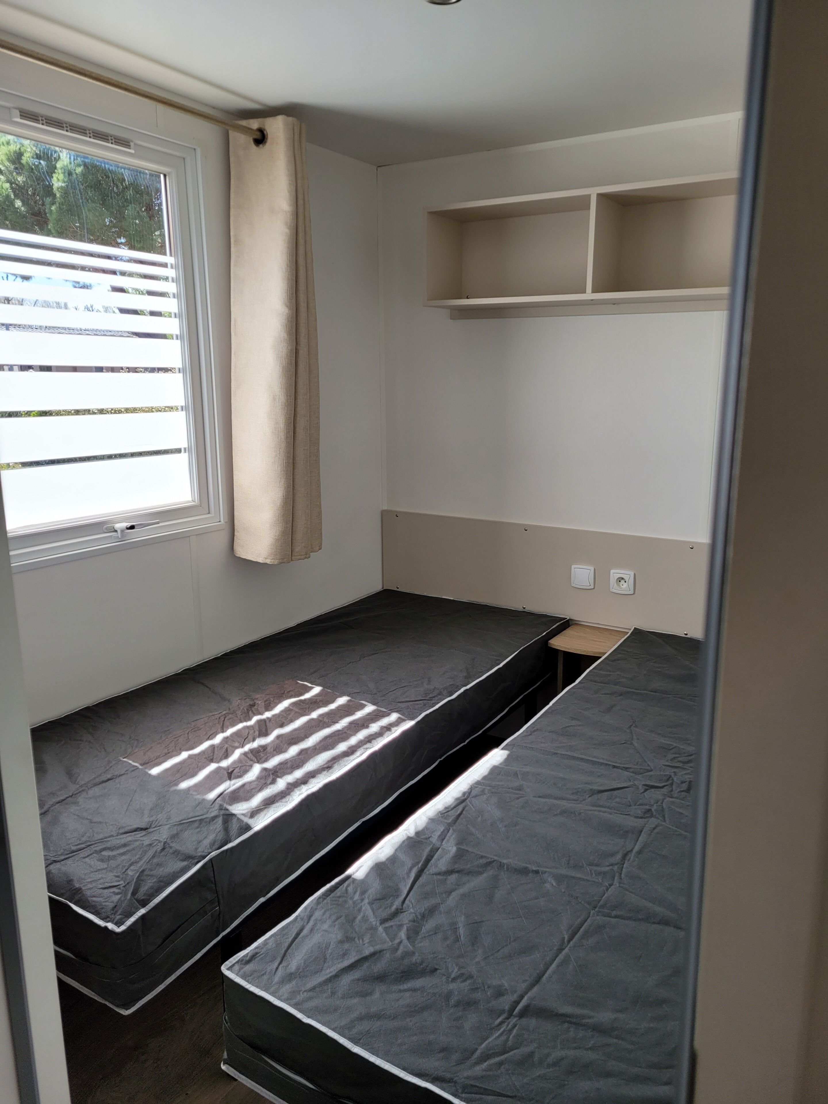 A compact room with a single bed, a small window with curtains, and a shelf unit.
