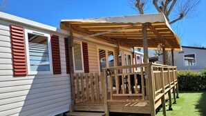 A wooden deck with steps leading to a covered porch, a white mobile home with red shutters, and a green lawn. - Vias Plage,la Carabasse Mobil Home Tout Confort (Vias-plage)