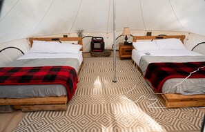 A tent with two wooden beds, each with a red and gray blanket, a heater, and a lamp on a wooden nightstand.