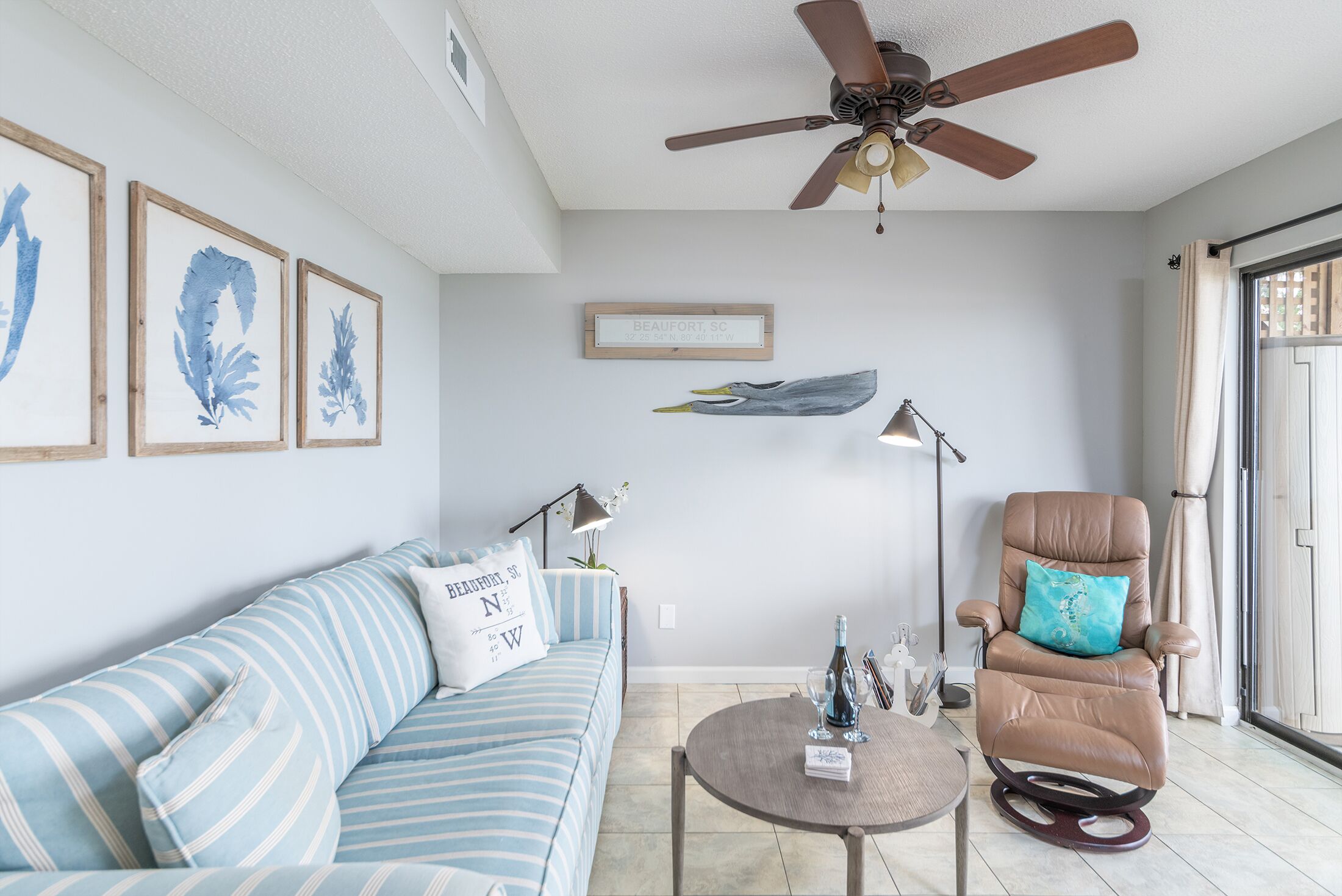 Un salon avec un canapé à rayures, un fauteuil inclinable, un ventilateur de plafond et des œuvres d’art encadrées accrochées au mur.