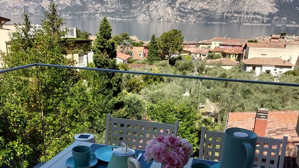 Een tafel voor twee, met bijpassend turquoise servies en een vaas vol roze bloemen, met uitzicht op een bergachtig landschap en een meer.