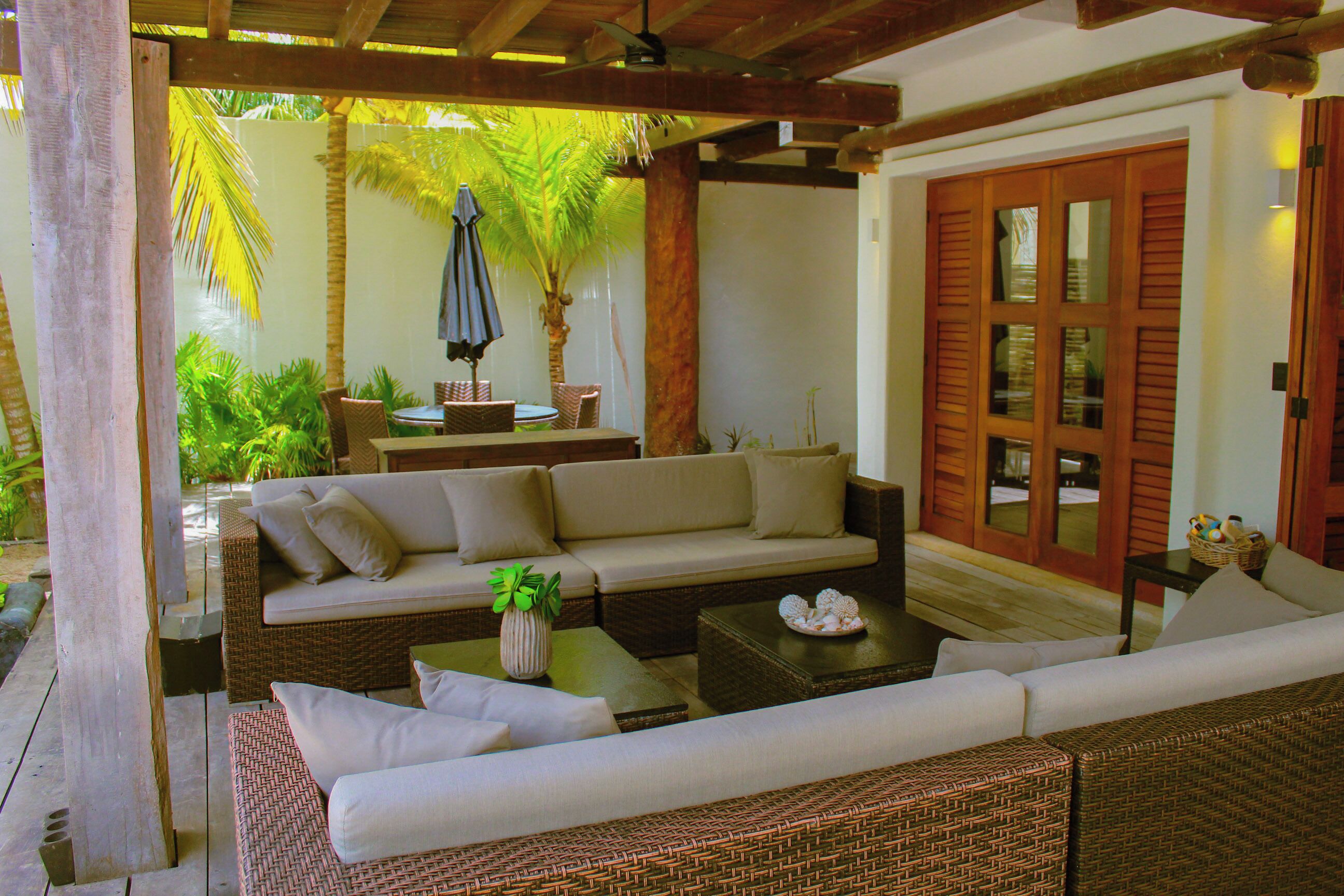 A covered outdoor seating area with wicker furniture, a palm tree, and a wooden door.