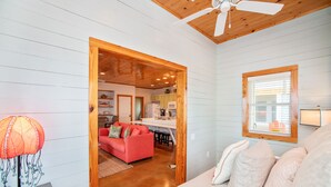 A cozy room with a wooden ceiling, a bed with pillows, a red sofa, and a kitchen area visible through an open doorway.
