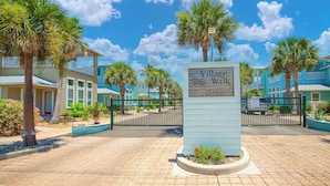 A sign for Village Walk, a residential area with a paved entrance, palm trees, and a clear blue sky.