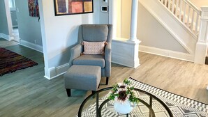 Un salón con un sillón gris, un tapete estampado, una mesa de centro con un centro de mesa floral, una escalera con barandillas blancas y un cuadro abstracto de gran tamaño en la pared.