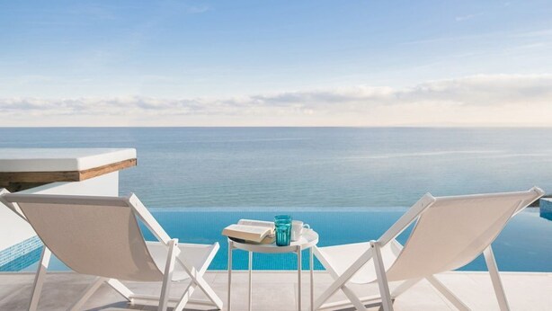 A poolside view with two white lounge chairs, a small table with a book and a glass, and a clear view of the sea. - Luxury fiore suites 3 (Zakinthos)