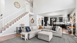 A spacious living room with a large sectional sofa, a white staircase, and a kitchen in the background.