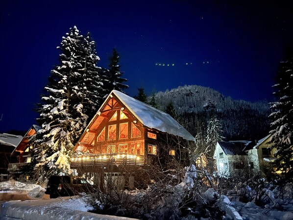 A log cabin with a snow-covered roof, surrounded by snow-laden trees and a mountainous backdrop.