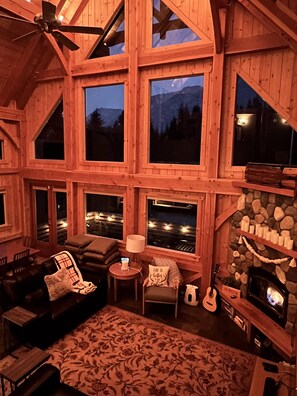 A cozy cabin interior with a fireplace, large windows, and a ceiling fan.