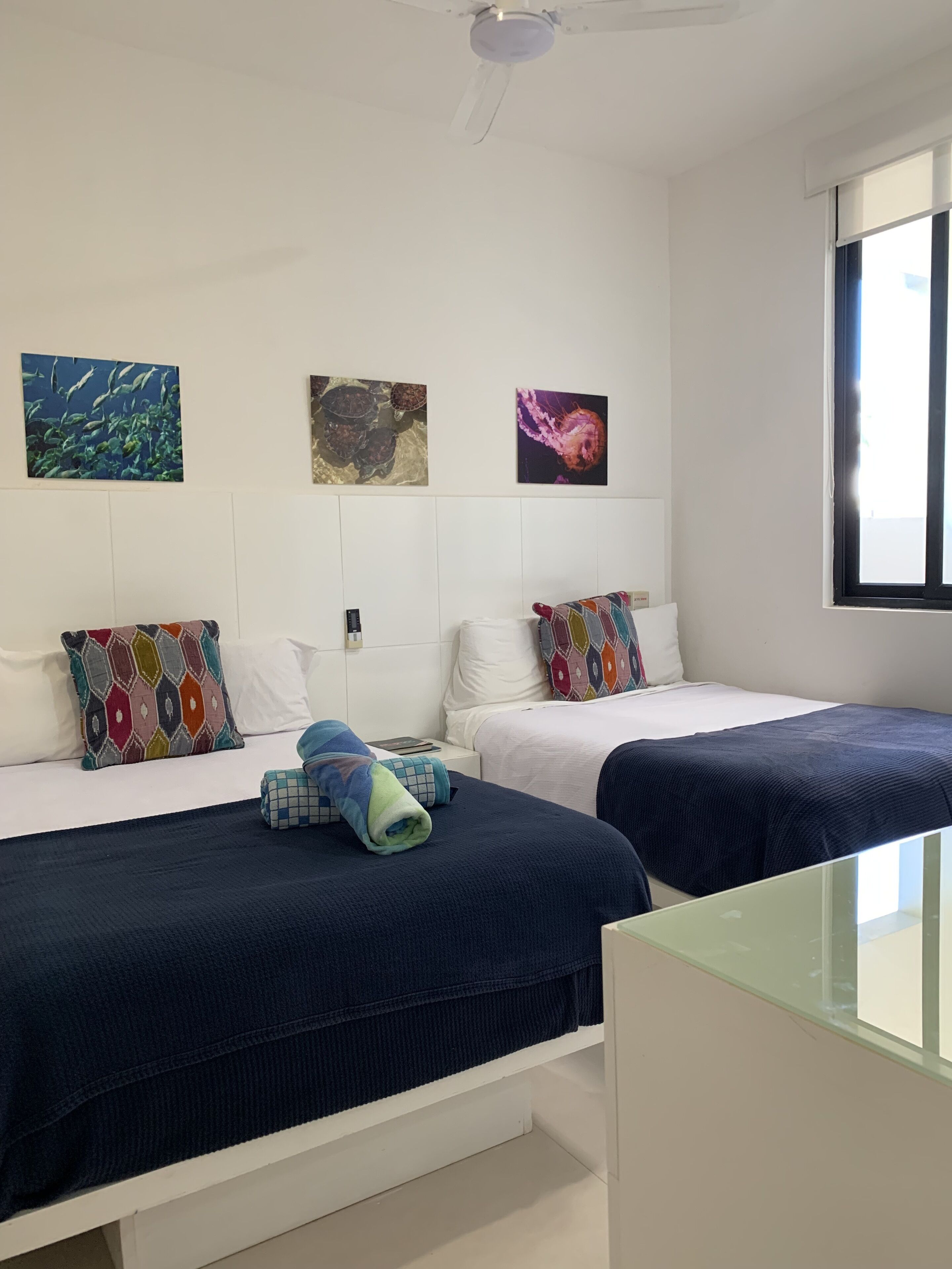 A hotel room with two beds, each with a colorful pillow, a ceiling fan, and artwork on the wall.