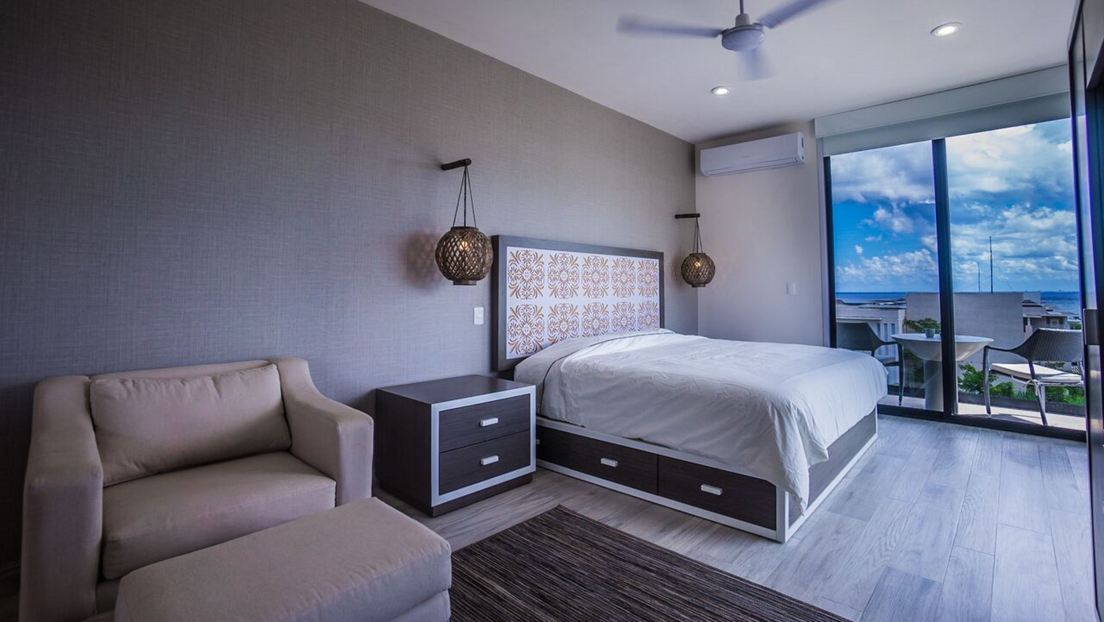 A modern bedroom with a large bed, a nightstand, a chair, and a balcony with a view of the ocean.