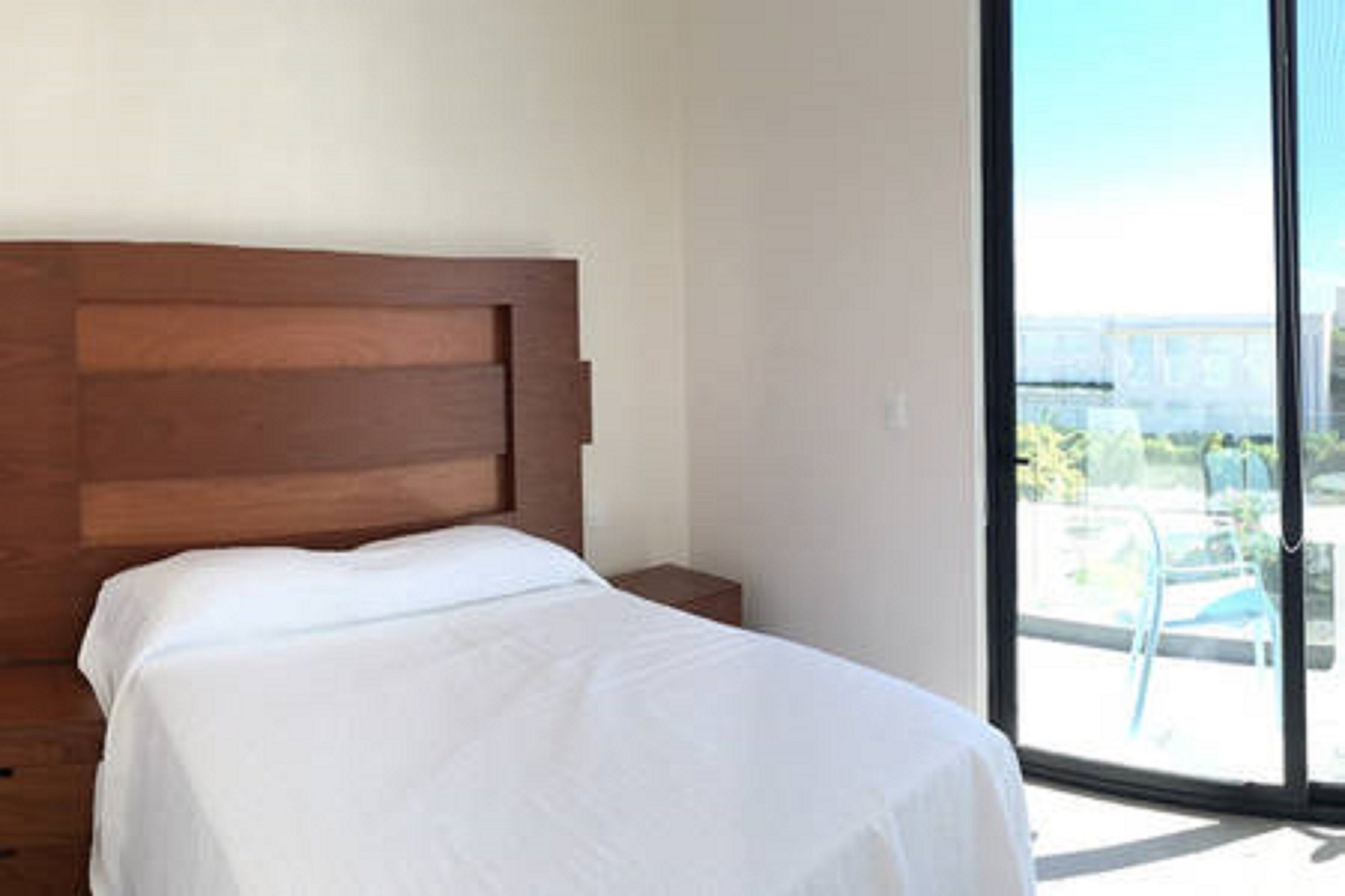 A hotel room with a large bed, a wooden headboard, a small bedside table, and a large window offering a view of the city.