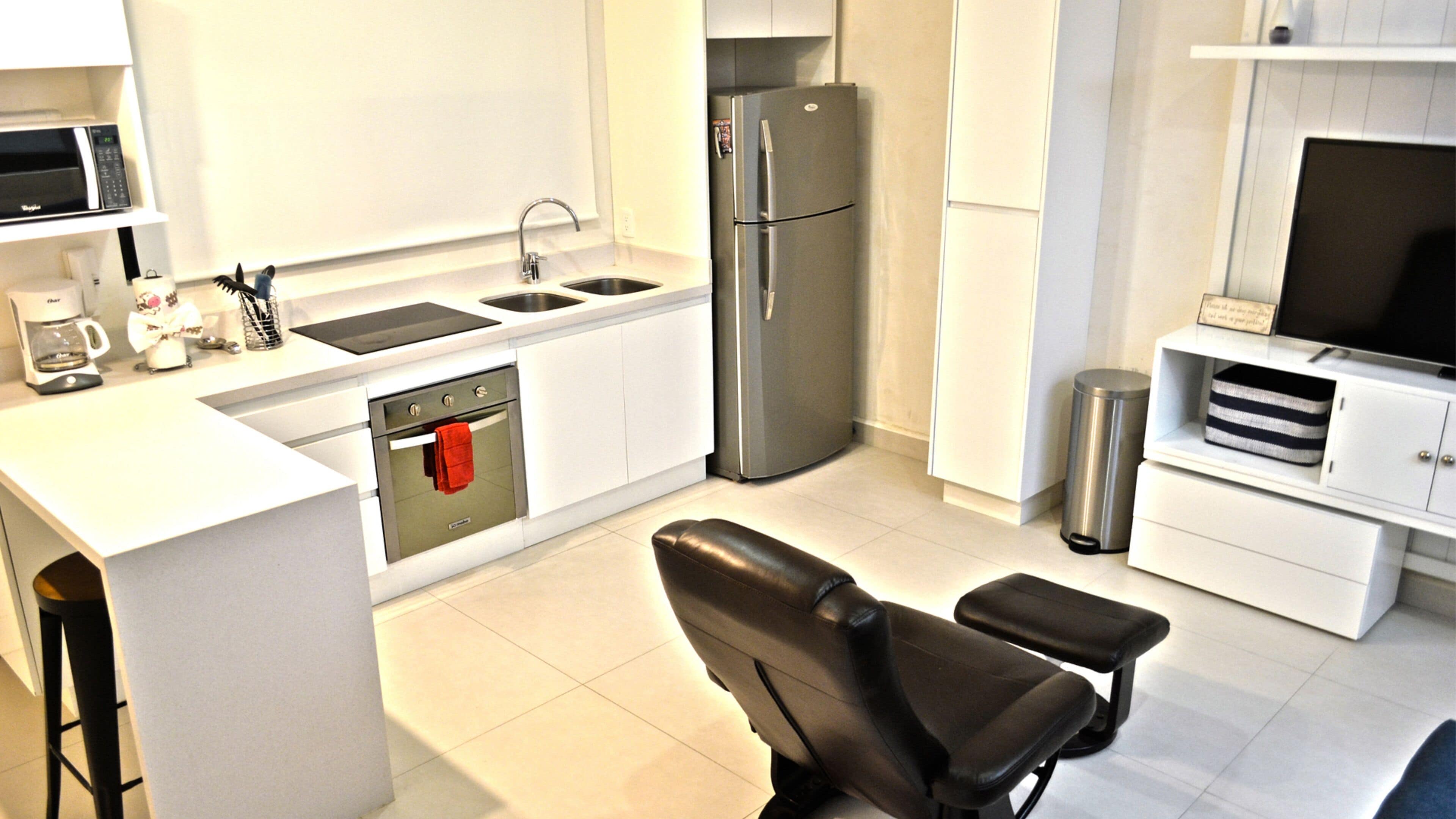Una cocina moderna con un refrigerador de acero inoxidable, un horno empotrado, un lavamanos y una cafetera. Hay una silla de cuero negro y un televisor sobre un mueble blanco.