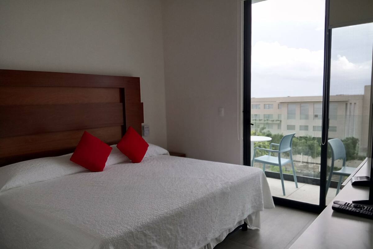 A hotel room with a bed, a balcony with chairs, and a view of buildings.