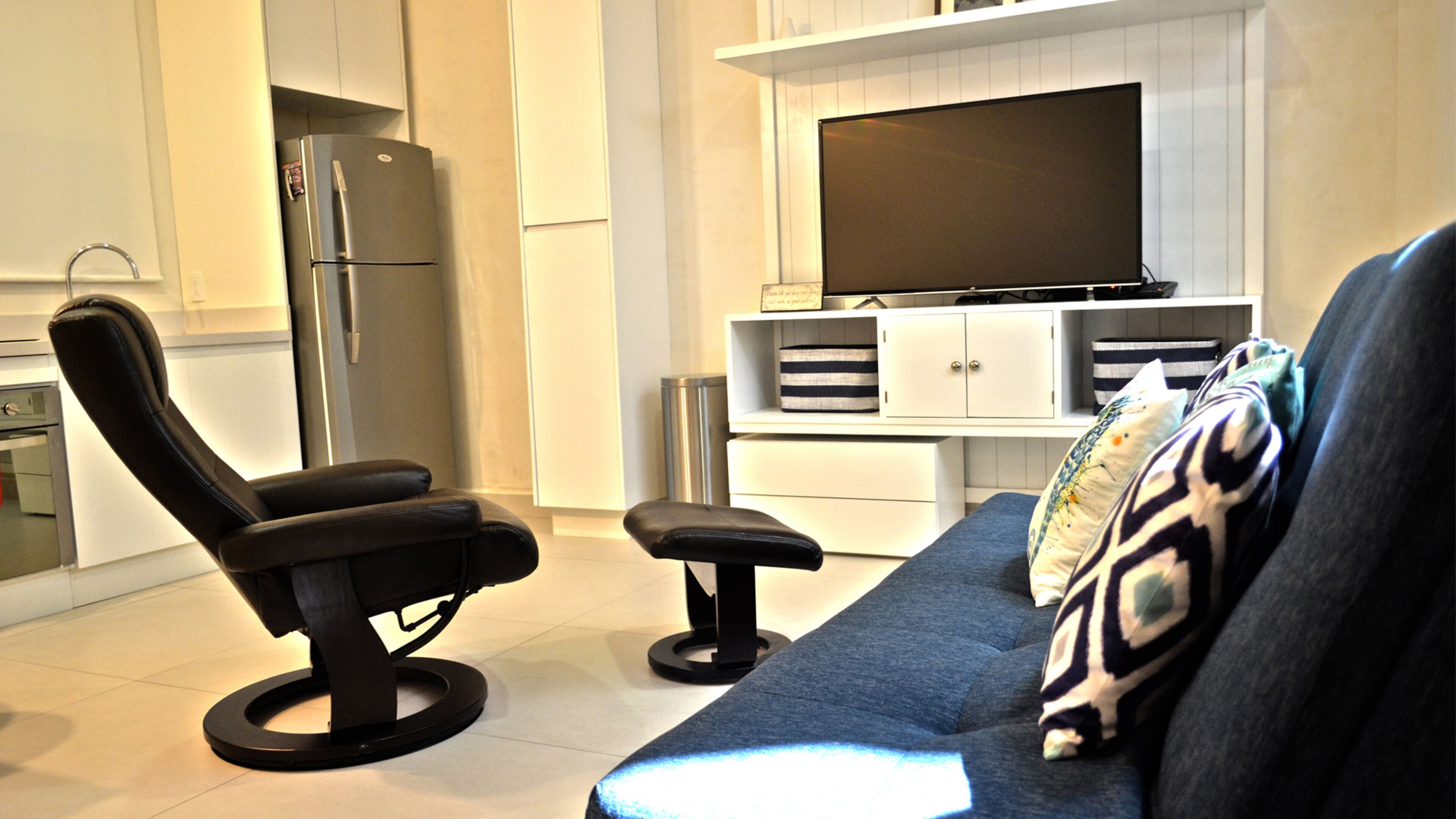 A modern living room with a black leather chair, a blue sofa, and a flat-screen TV mounted on a white cabinet.