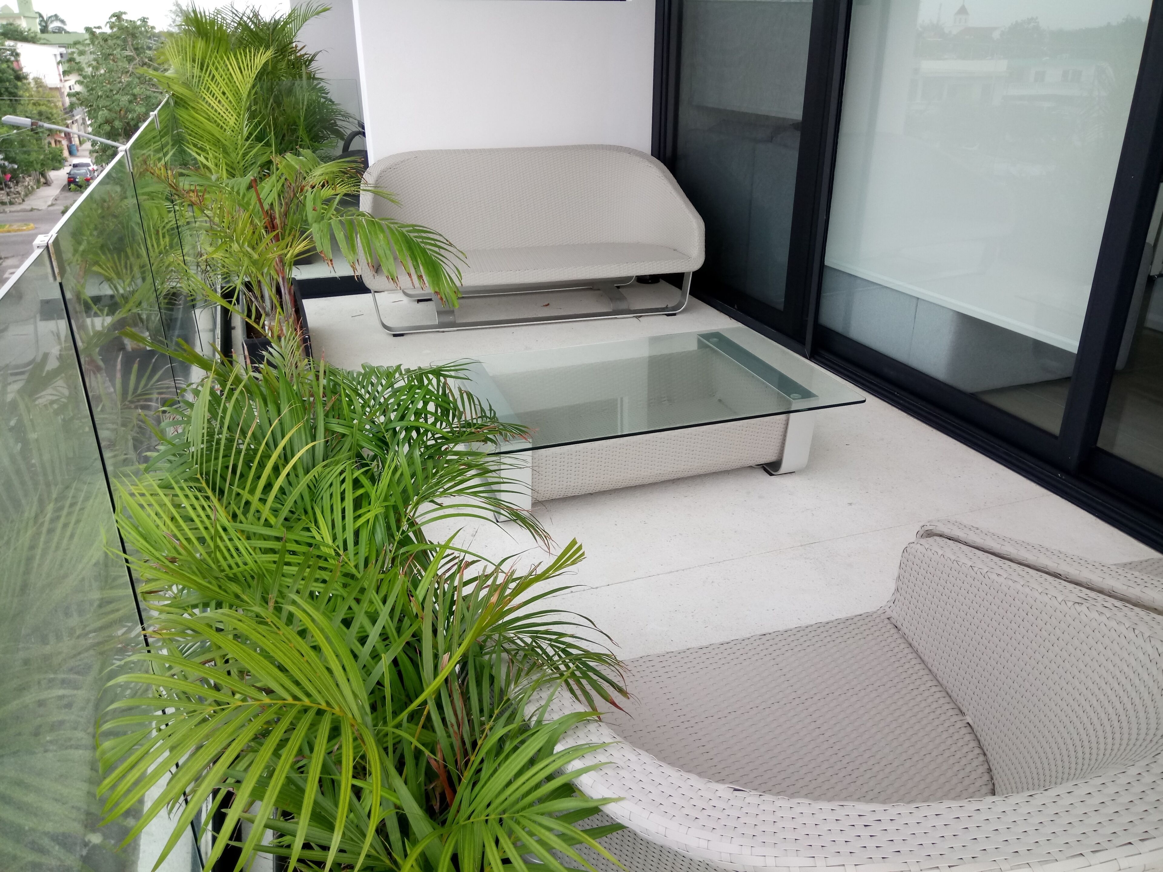 A modern balcony with a glass table, a white sofa, and lush green plants.