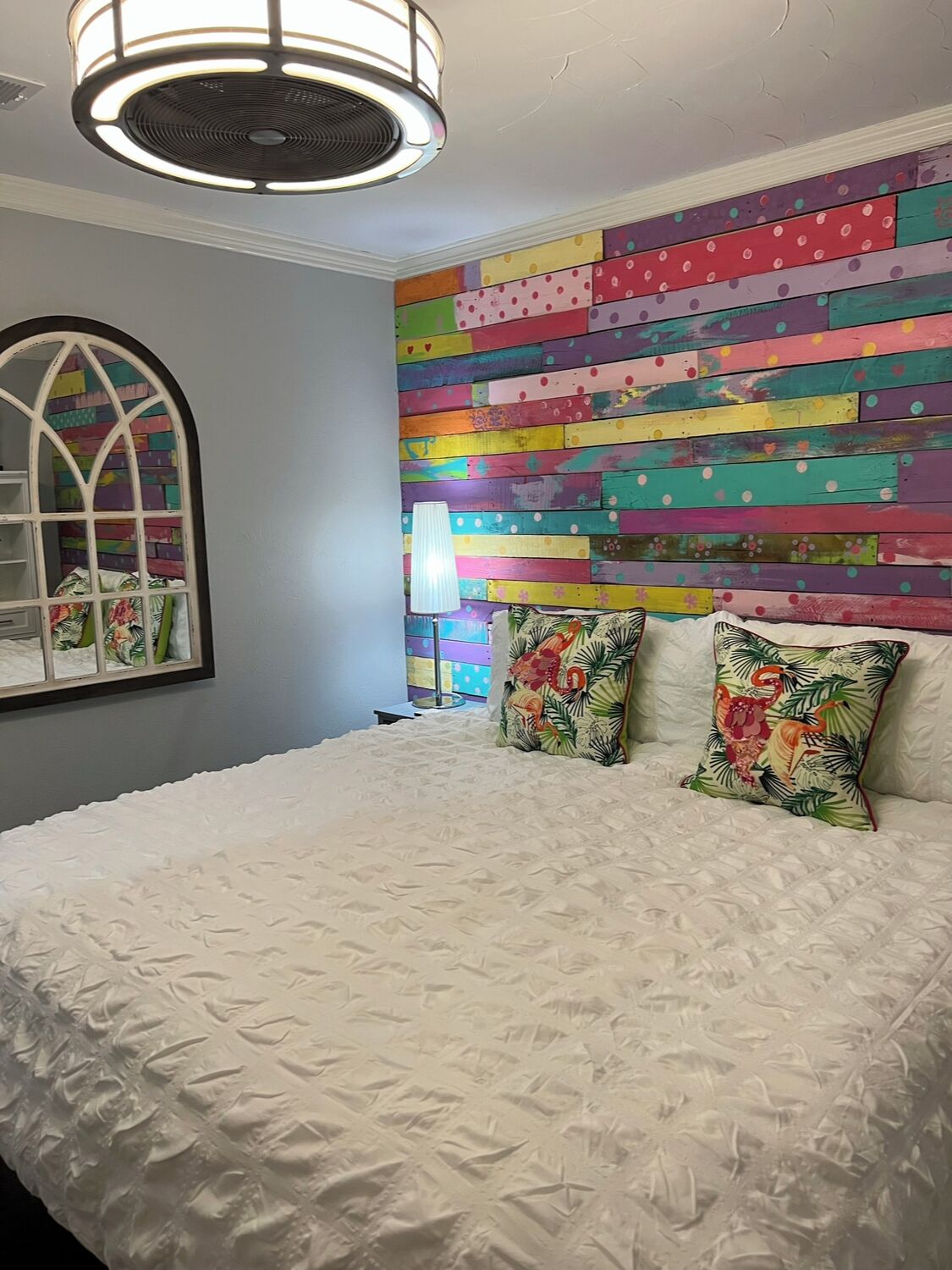 A bedroom with a large bed, colorful wooden wall paneling, a decorative mirror, and a ceiling fan.