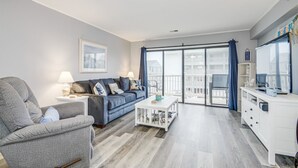 Ett modernt vardagsrum med en grå soffa, ett vitt soffbord och en grå fåtölj. Det finns ett stort fönster med utsikt över en balkong och byggnader.