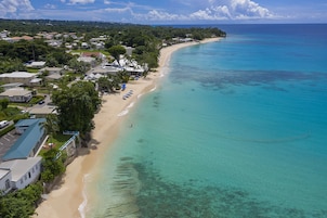 An aerial view of a coastal area with a sandy beach, clear turquoise waters, and residential buildings along the shore.