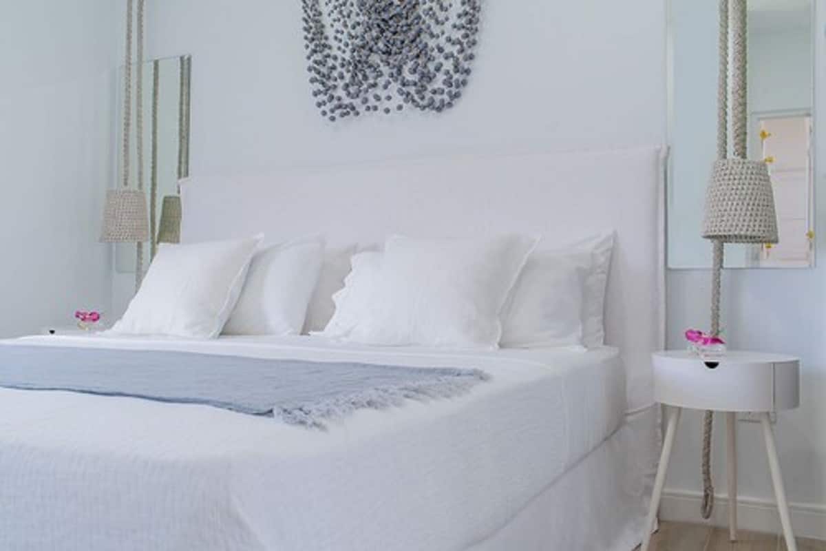 A neatly made bed with white linens and pillows, a bedside table with a lamp, and a wall-mounted mirror.