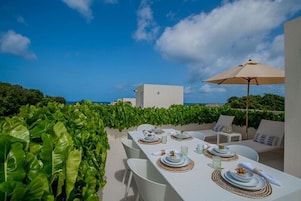 An outdoor dining area with white tables and chairs, a large umbrella, and a view of the ocean.