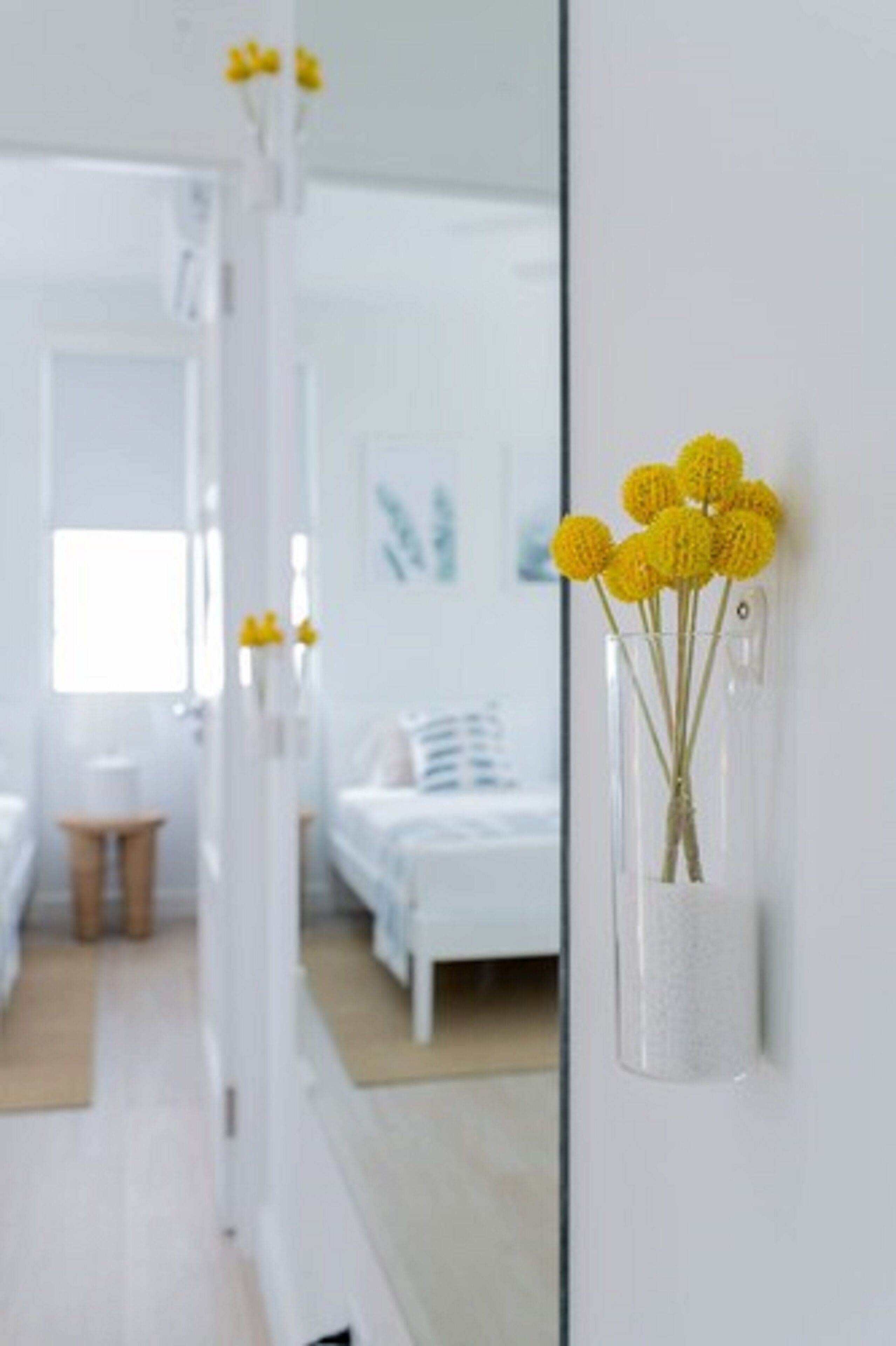 A modern hotel room with a large mirror reflecting the interior, a white bed, and a vase with yellow flowers on the wall.