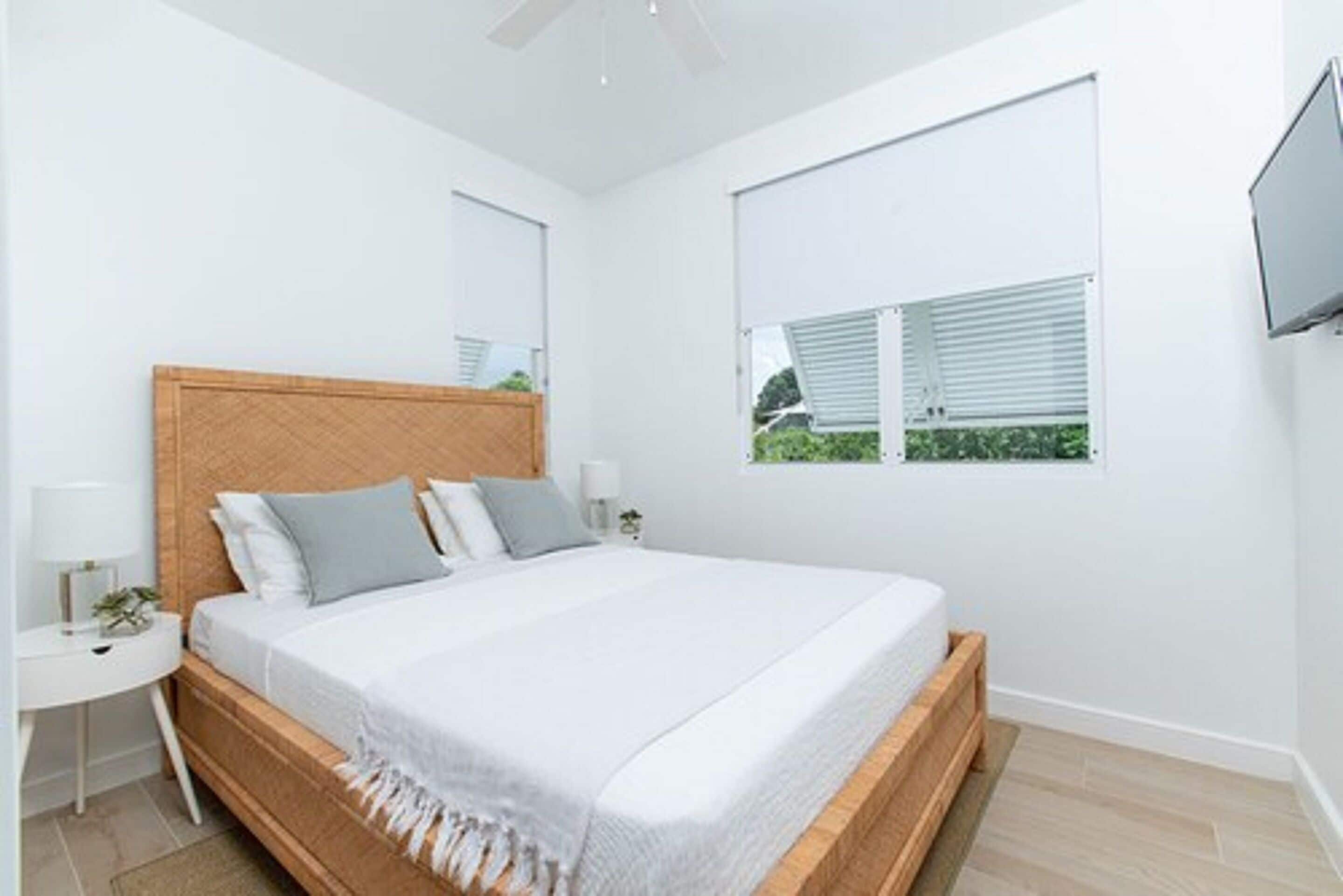 A modern bedroom with a wooden bed frame, white bedding, a bedside table with a lamp, and a window with blinds.