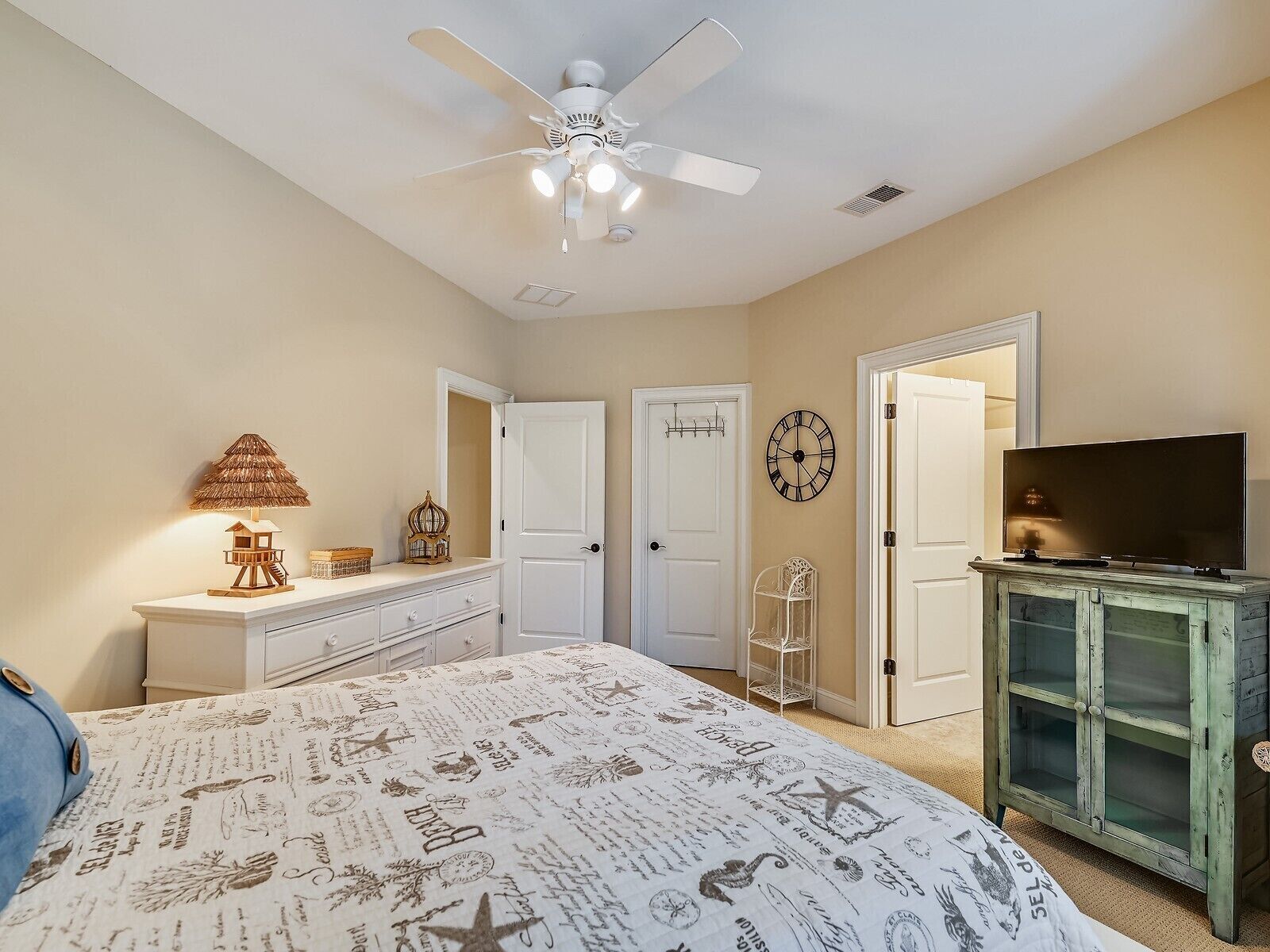 A bedroom with a bed, dresser, vanity, mirror, clock, television, and a ceiling fan.
