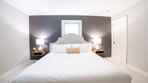 Un grand lit avec une tête de lit blanche, deux tables de chevet avec des lampes, une fenêtre et une porte.