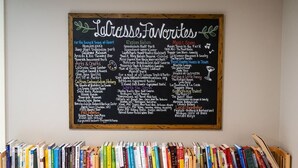 Un tableau noir présente différentes catégories de lieux et d’activités prisés à LaGrasse, notamment « Pour les jeunes et les jeunes d’esprit », « À la découverte de la nature », « Arts et artisanat » et « Scène musicale ». En dessous du tableau noir, on trouve une bibliothèque remplie de divers ouvrages.