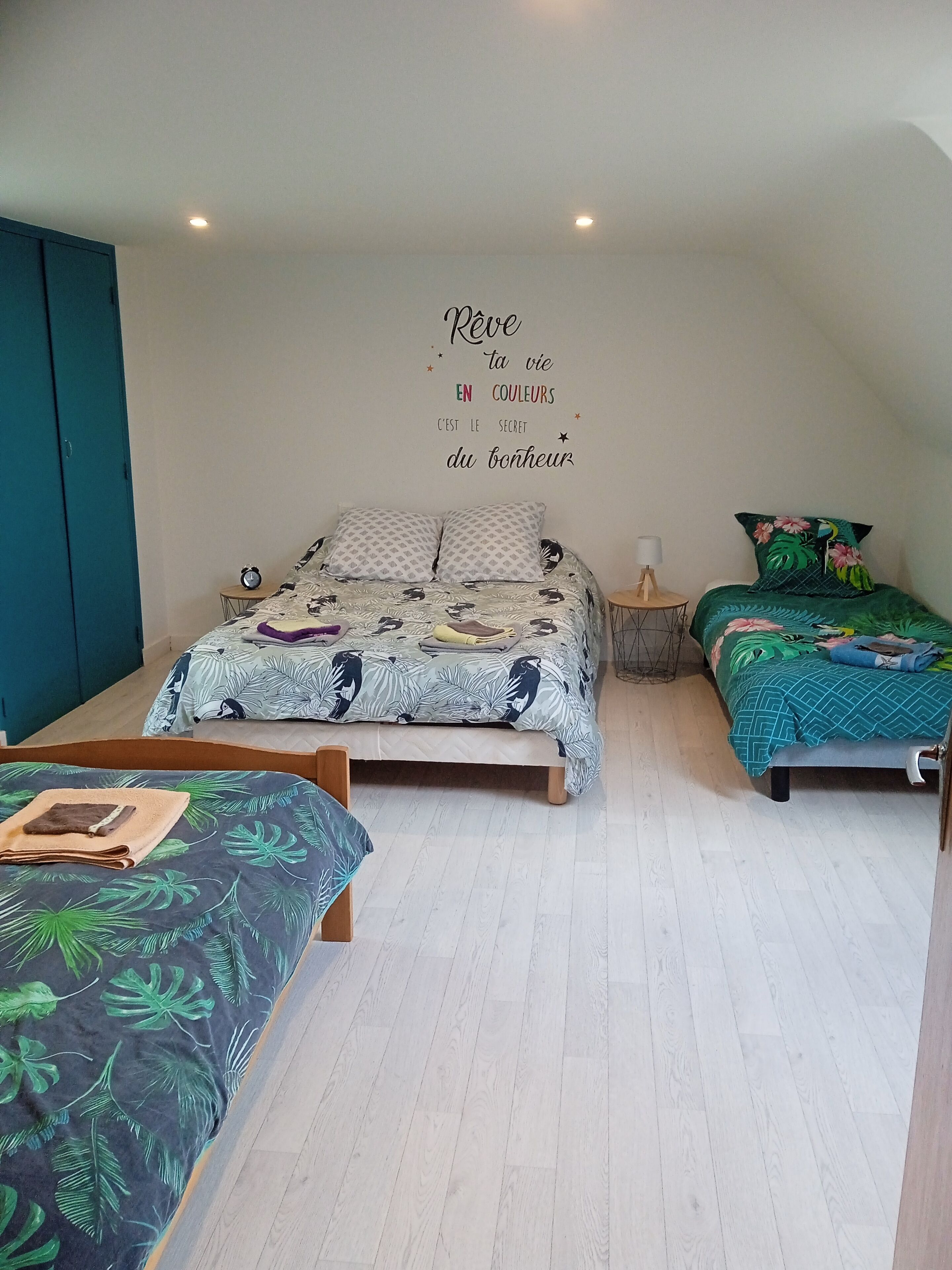 A bedroom with a large bed, two smaller beds, a wall clock, a bedside table with a lamp, and a wall with a motivational quote.
