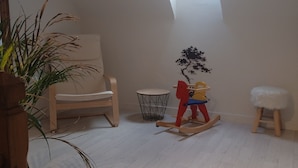 A room with a sloped ceiling, a small window, a rocking horse, a chair, a stool, and a plant. - Grande Longère (ploezal)