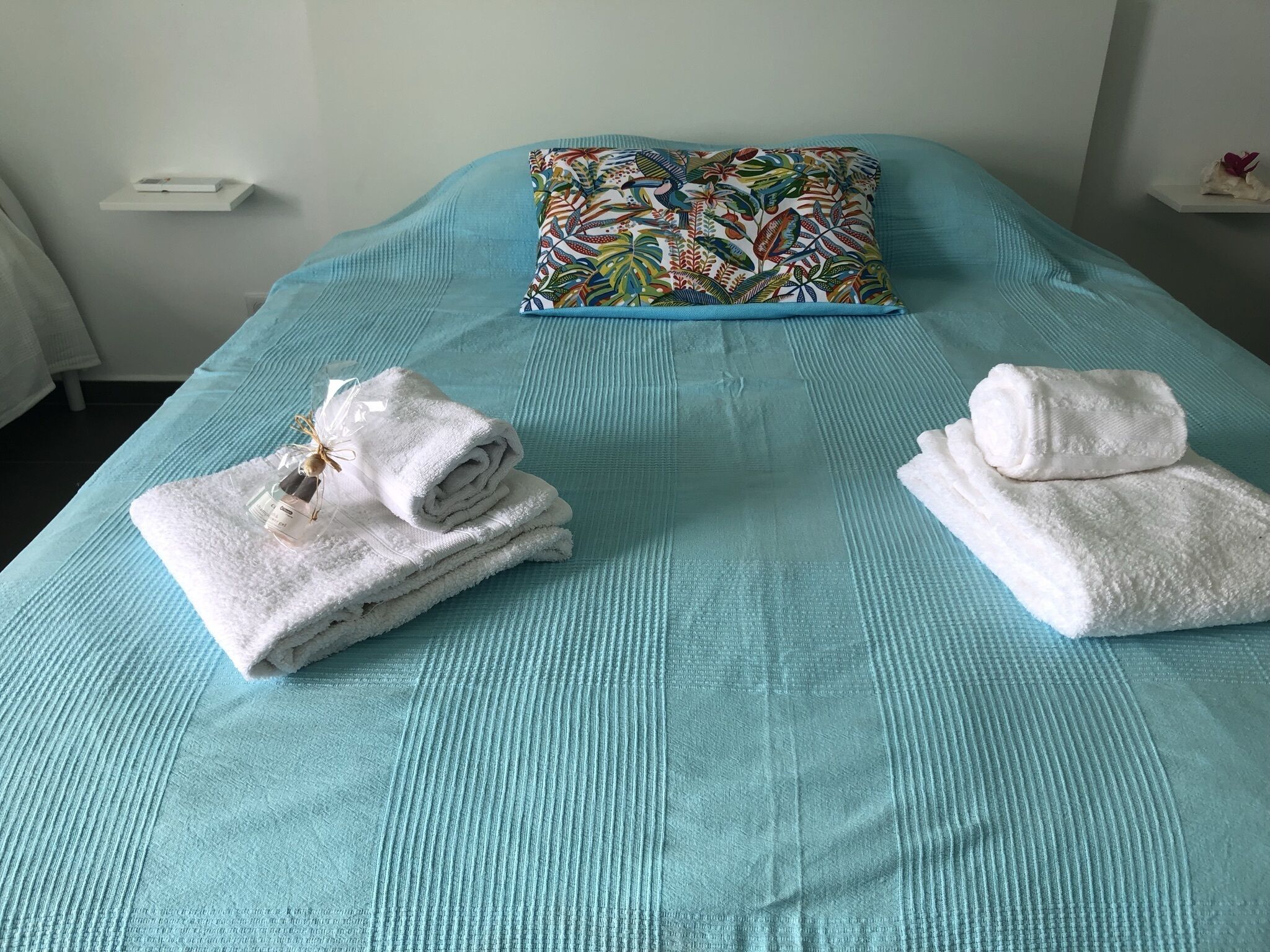 Un lit bien fait, recouvert d’une courtepointe turquoise, agrémenté d’un coussin aux motifs floraux colorés et de deux serviettes blanches soigneusement pliées, avec un petit flacon posé sur l’une d’elles.