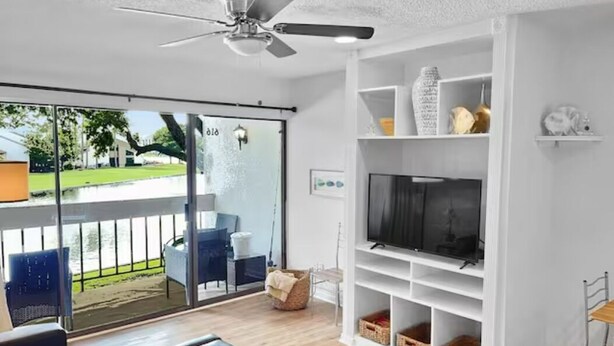 Un salón moderno con un televisor grande, estanterías blancas y un balcón con mobiliario exterior.