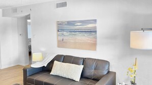 Una sala de estar moderna con un sofá de cuero, una mesa de centro con controles remotos, un cuadro de una playa, una lámpara y una mesita con un cartel de 'Welcome' y flores.